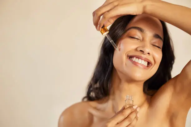 Happy young woman with bare shoulder applying serum on face with closed eyes. Beautiful hispanic young woman moisturizes her skin with serum isolated against beige background with copy space. Healthy and skin care conscious girl using facial treatment in summer.