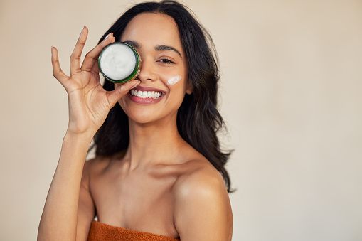 Happy attractive young woman hold green cream jar and covering eye while looking at camera with a big laugh. Portrait of multiethnic girl with bare shoulder showing natural organic cream box isolated against beige background with copy space. Mixed race beauty woman with organic and herbal cream for healthy and smooth skin.