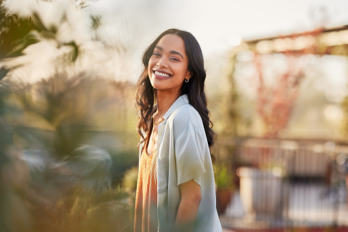 Young latin woman in casual clothing in the garden looking at camera, during early morning. Portrait of healthy mexican girl enjoying nature during sunset. Mindful multiethnic woman enjoy morning ritual with fresh air.