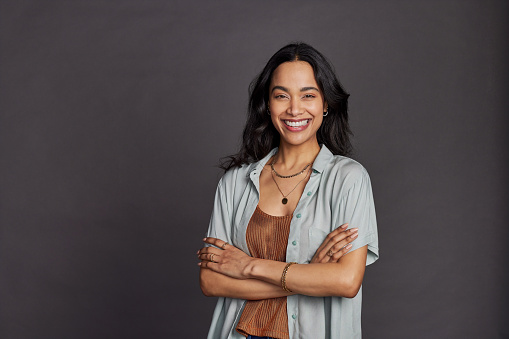 Portrait of smiling young multiethnic woman in casual clothing with arms crossed standing against grey background. Cheerful latin woman with folded hands looking at camera with big grin. Satisfied mexican girl with copy space isolated against gray background.