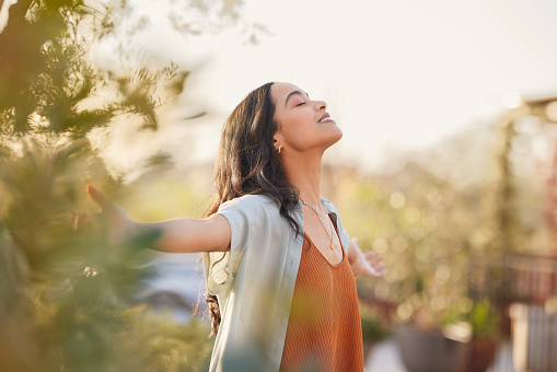 Young latin woman with arms outstretched breathing in fresh air during sunrise at the balcony. Healthy girl enjoying nature while meditating during morning with open arms and closed eyes. Mindful woman enjoying morning ritual while relaxing in outdoor park.