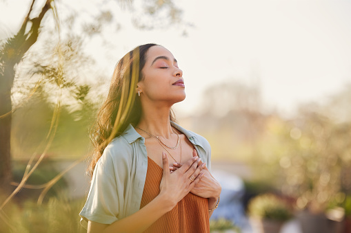Young latin woman with hand on chest breathing in fresh air in a beautiful garden during sunset. Healthy mexican girl enjoying nature while meditating during morning exercise routine with closed eyes. Mindfulness woman enjoying morning ritual and relaxing technique.