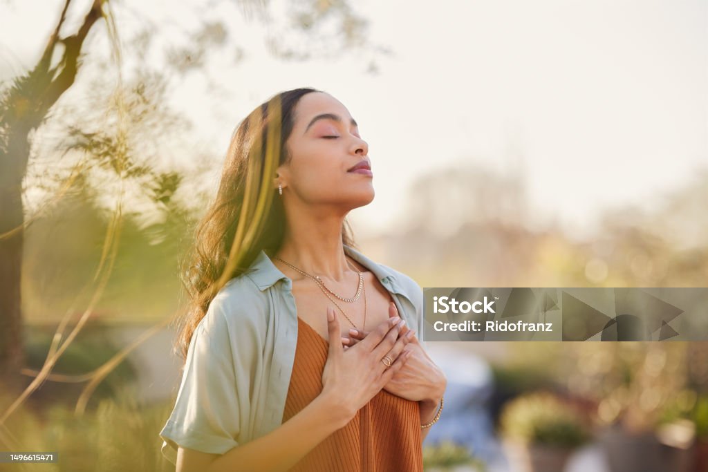 Mixed race woman relax and breathing fresh air outdoor at sunset Young latin woman with hand on chest breathing in fresh air in a beautiful garden during sunset. Healthy mexican girl enjoying nature while meditating during morning exercise routine with closed eyes. Mindfulness woman enjoying morning ritual and relaxing technique. Gratitude Stock Photo
