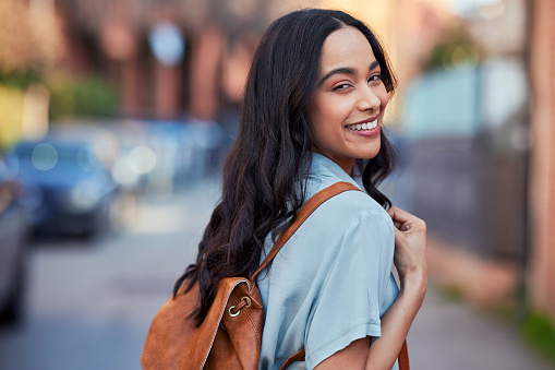 Portrait of smiling young multiethnic woman with back pack moving freely in city streets. Mixed race girl traveling in city during vacation. Latin carefree student going to university lesson while looking at camera.