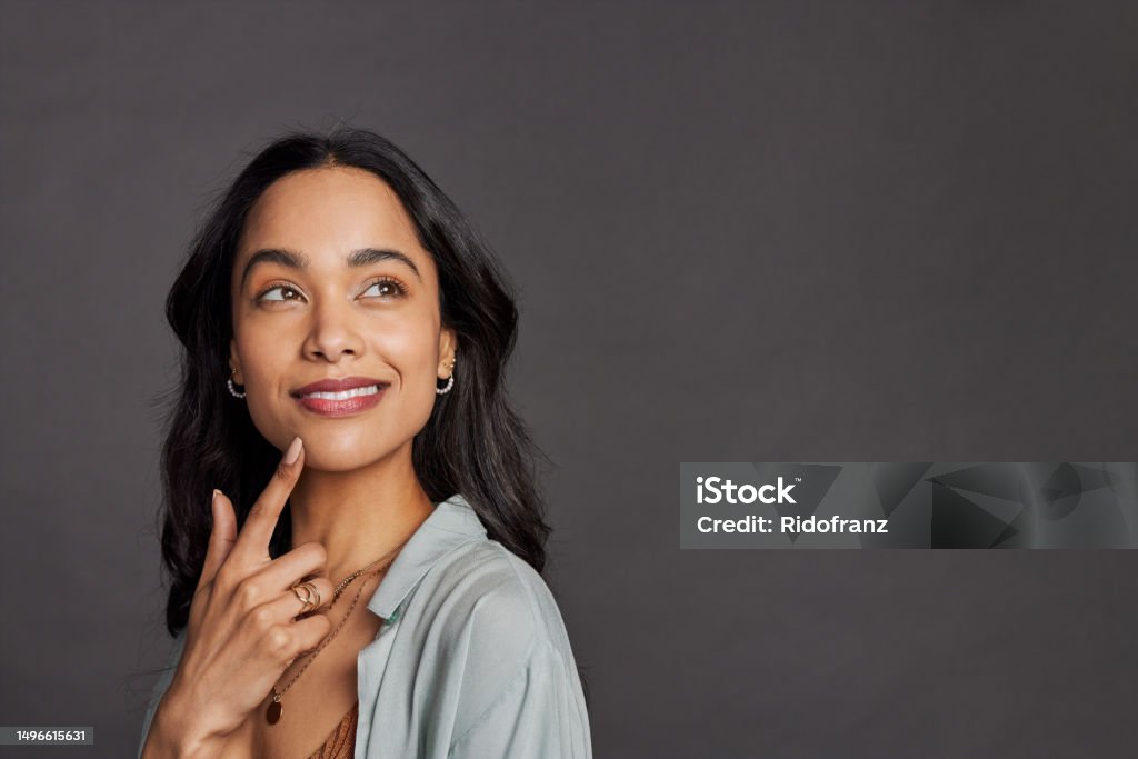 Pensive young multiethnic woman looking up on grey wall Portrait of pensive young mixed race woman with hand on chin looking away isolated on gray background. Multiethnic beautiful girl in casual clothing isolated against grey wall daydreaming with copy space. Close up face of happy mexican woman thinking and making plans about future. Women Stock Photo