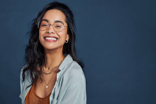 Cheerful young woman with eyeglasses smiling and looking at camera Casual young multiethnic woman with eyeglasses smiling at camera on blue background. Close up face of happy mixed race girl laughing with eyeglasses isolated with copy space. Beautiful university student having fun isolated against blue wall while wearing specs. brazilian ethnicity photos stock pictures, royalty-free photos & images