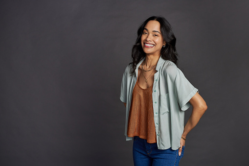 Portrait of smiling young woman in casual clothing standing against gray background. Cheerful multiethnic girl with hands in jeans back pockets looking at camera while smiling. Young mixed race woman looking at camera with copy space isolated against grey wall.