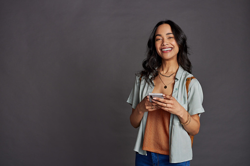 Happy casual beautiful woman using smartphone isolated against grey background. Smiling hispanic young woman with backpack using mobile phone isolated against gray background with copy space. Mixed race girl subscribing to new social media channel on smart phone.