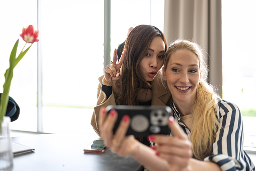 Dark haired mystical lady of Asian European origin in a black dress is smiling and hanging out with her blonde friend in a cafe, they are scolding each other and taking selfies with a mobile phone.