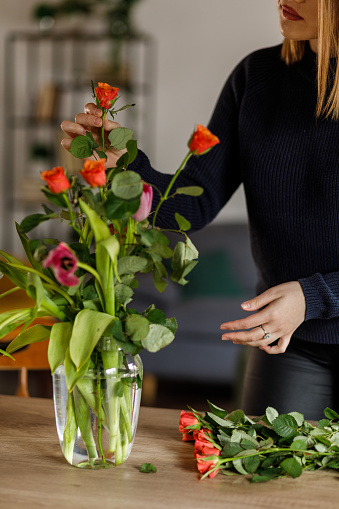Close up shot of diligent young woman bringing spring in her home by arranging roses and tulips in a bouquet in a glass vase at her dining table.