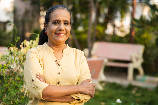 Happy smiling indian senior woman confidently standing with arms crossed by looking camera at park - concept of retirement lifestyle , successful and relaxation.