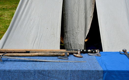 A close up on several medieval axes, maces and swords displayed on a blue cloth next to a white cloth tent stretched by means of ropess seen on a sunny summer day on a Polish countryside during a hike