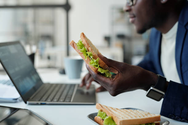 Focus on appetizing vegetarian sandwich in hand of young hungry African American manager in formalwear using laptop at lunch break