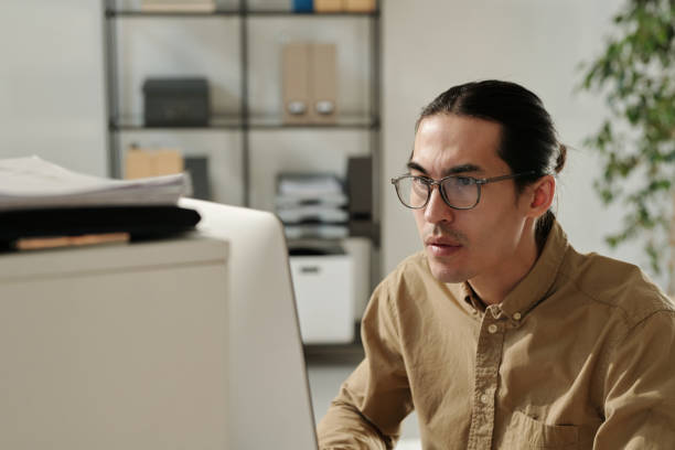 istock Young serious analyst in eyeglasses sitting by workplace in front of computer 1496639175