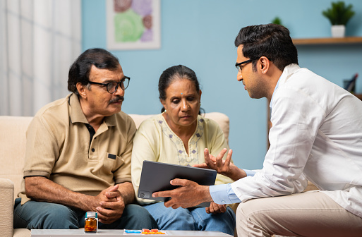 Indian doctor explaining from digital tablet to senior couple about medical report at home - concept of technology, health care support and home consultation