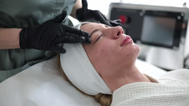 Pretty girl receiving cosmetic facial care at a spa salon. A cosmetologist is performing a therapeutic procedure on her face. Cosmetologist gently applies scrub massages the face with a gel mask.