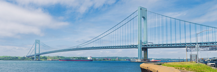 High resolution stitched panorama of the Verrazano-Narrows Bridge on a Cloudy Summer Morning with Promenade, Cars Leaving the Bridge and Driving on Belt Parkway and Cargo Ship. The bridge connects boroughs of Brooklyn and Staten Island in New York City. The bridge was built in 1964 and is the largest suspension bridge in the USA. Historic Fort Wadsworth is under the bridge. The photo was taken from the Shore promenade in Bay Ridge Brooklyn. Canon EOS 6D full frame censor camera. Canon EF 85mm F/1.8 lens. 3:1 Image Aspect Ratio. This image was downsized to 50MP. Original image resolution is 95.2MP or 16896 x 5632 px.