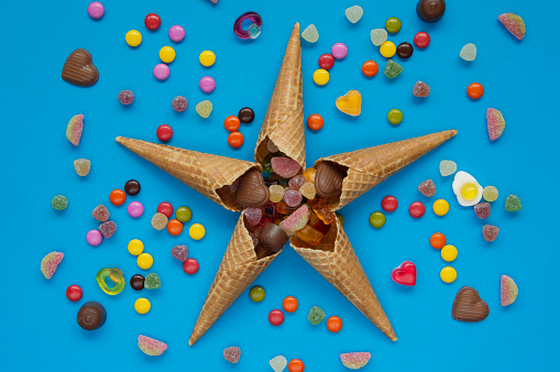 Candy splash out of the sweet cones in the star shape. Candy mix with jellybeans, gummy candy, mint, cone, sweets and bonbons on blue background.