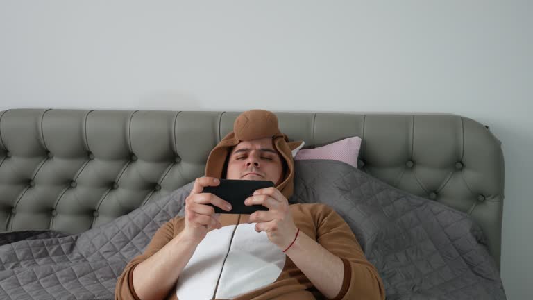 Man in cosplay costume of a cow. Guy in the funny animal pyjamas lie on the bed and playing games on mobile phone. Weekend, relax at home. Guy chatting in social network.