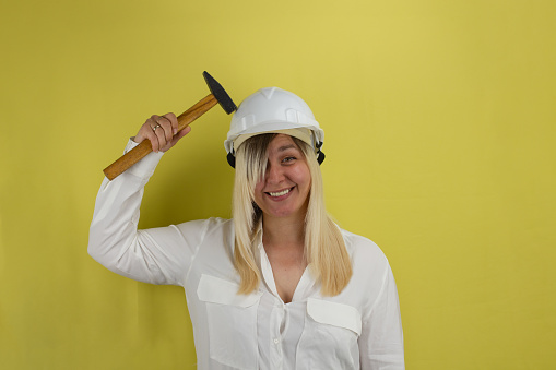 On a yellow background, a cute woman with a silly grin hits the helmet on her head with a hammer and smiles. Funny woman with a hammer and in a construction helmet on a yellow background isolated..