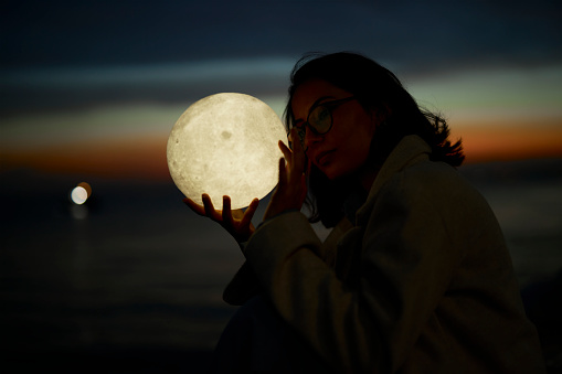 beautiful girl with fashionable dress enjoying blue hours through sunset near seaside with a led lamp in moon shape