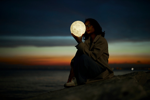 beautiful girl with fashionable dress enjoying blue hours through sunset near seaside with a led lamp in moon shape