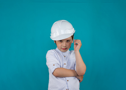The boy on a blue background is isolated in a construction helmet and a white shirt. A handsome boy in the role of a builder looks at the camera. The child imagines himself as a builder in the future.