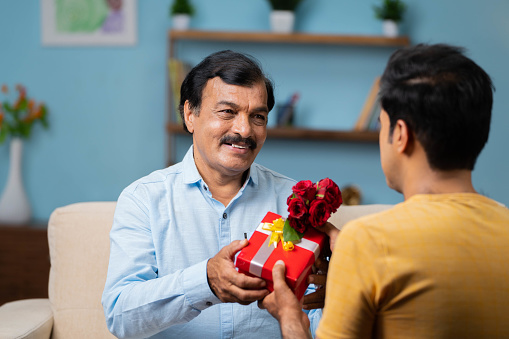 Happy senior father receiving gift with flowers from son at home - concept of father day celebration, family bonding and relationship.