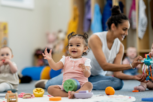 A small group of babies sit on the floor playing with various toys while their teacher supervises and engages them.  They are each dressed casually  and appear content.