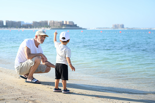 Adorable two year old baby boy throwing rocks sand seashells in the sea and his grandpa playing on the beach