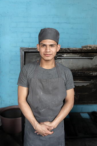 Portrait of a young Hispanic man posing in front of the bakery oven with a blackuniform. Concept of traditional Mexican sweet bread craft