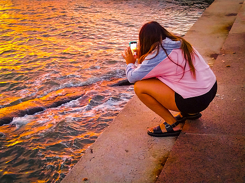 Teen woman crouched taking photos of sunset at coast beach, montevideo, uruguay