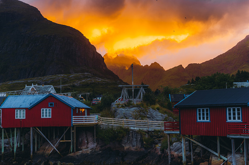 August 4th, 2022: Dramatic view of the bright colorful sky above the mountains and the old fisherman village Å with red houses and reflection sea during the Midnight Sun on Lofoten, Northern Norway