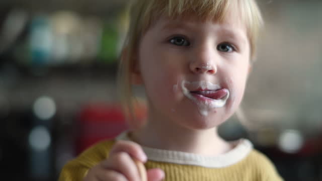 Little girl is learning to use spoon while eating yoghurt at home in the kitchen