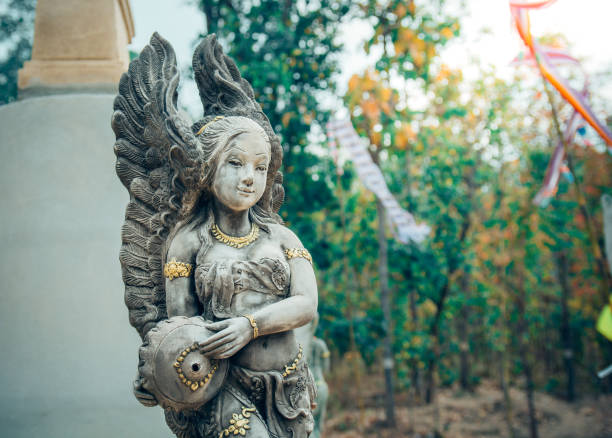 Female statue with angle like wings among trees at temple Wat Phrathat Doi Wiang Chai Mongkhon วัดพระธาตุดอยเวียงชัยมงคล – Temple Thai Ping Khong, Chiang Dao District, Chiang Mai. Temple is ancient one from early time of Chiang Mai kingdom more than 700 years ago. Behind statue amd tree is another femlae statue nwith hands folded in Wai gesture