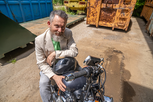 An elderly man wearing a light green t-shirt, beige jacket, and gray jeans with a Bobber motorcycle. He is sitting on the bike and resting with his hands on the helmet. Wide angle view.