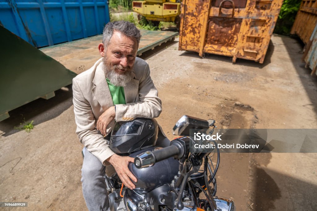 A mature man wearing a light green t-shirt, a beige jacket, and gray jeans on a Bobber Motorcycle. An elderly man wearing a light green t-shirt, beige jacket, and gray jeans with a Bobber motorcycle. He is sitting on the bike and resting with his hands on the helmet. Wide angle view. 50-54 Years Stock Photo