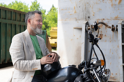 A mature man wearing a light green t-shirt, a beige jacket, and gray jeans on a Bobber Motorcycle. He has his hands resting on a black helmet. In the background - a green and rusted metal container.