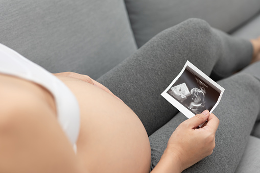 Close up Happy Pregnant Woman holding ultrasound image stroking big belly with love isolated on couch,Pregnancy of young woman enjoy with future life,Motherhood and Pregnant Concept