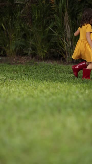 A Happy 4-Year-Old Cuban-American Toddler Girl with Brown Eyes & Brown Curly Hair Wearing a Yellow Cotton Dress & Shiny Red Rain Boots While Enjoying the Springtime Outdoors Running Around, Playing Kick Ball in Her Grassy South Florida Backyard, June 2023