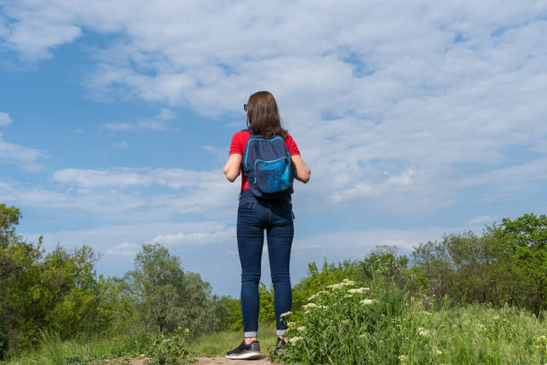 Tourist woman with backpack stand on edge and look on deciduous forest. Active adult leisure of hiking in green valley. Person enjoy wanderlust and view of the spring grassy hill. Female hiker explore.