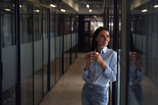 Dreamy young corporate worker with cup in her hands leaning against glass wall in corridor