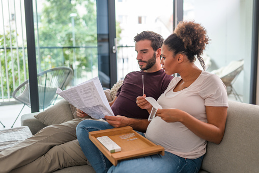 Side View Of A Young Pregnant Couple Sitting On The Sofa And Reading Instructions For Rapid Self Test