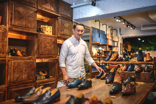 Waist up shot of a mid adult Chinese man enjoying a shopping day. He is looking for discounted leather shoes in a luxury footwear store. He is walking, wearing casual clothing and looking at the shoes on display.