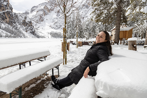 Woman smiling sitting leaning back and relaxing by Braies Lake completely iced in a snowy winter day; Alto-Adige, Italy; Dolomites