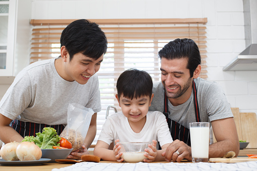 Happy cheerful gay couple with their adopted little son enjoy making a corn flakes with fresh milk for breakfast in the kitchen together. Happy LGBTQ+ family with adopted child concept.