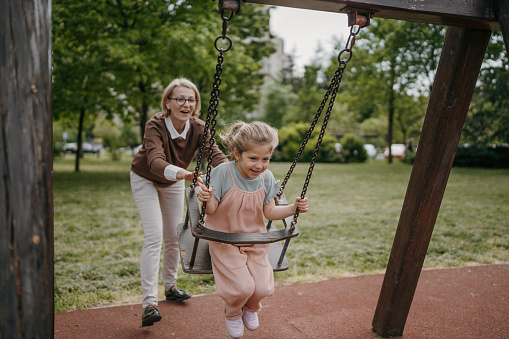 Young grandmother pushing her little granddaughter who is sitting on a swing in public park outdoors.