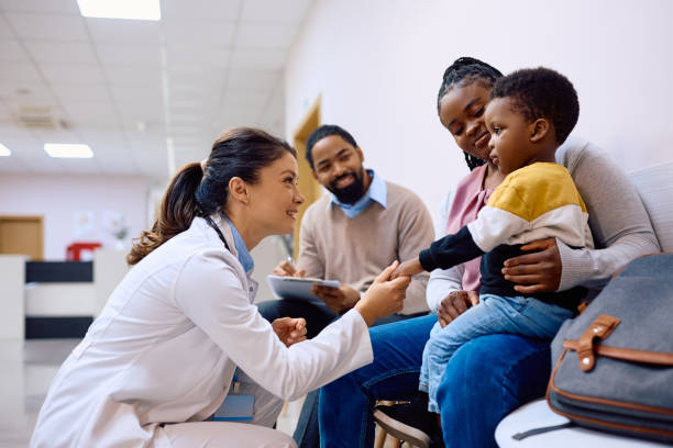 Female pediatrician talking to black little boy who is sitting on mother's lap at the clinic. Smiling doctor holding hand on African American boy who came with parents for medical exam at pediatric clinic. waiting room stock pictures, royalty-free photos & images
