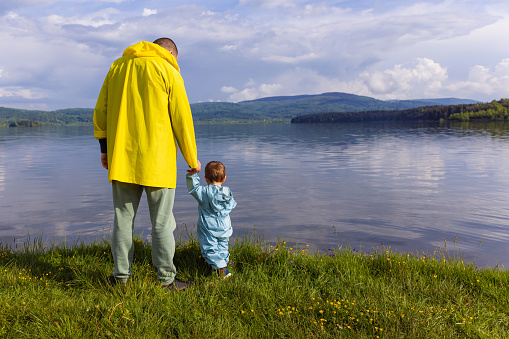 Father and his young son are wearing raincoats and standing by the lake, holding hands and enjoying nature.
