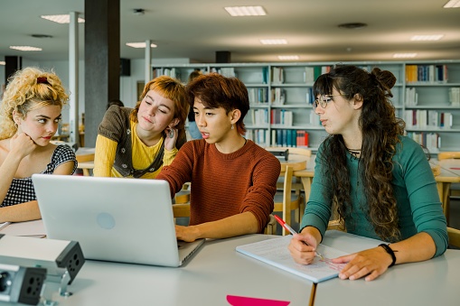 Group of multiracial student friends sitting at table in library looking at screen of laptop and Asian male explaining project while female blonde leaning on hand and looking away